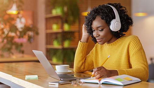 Smiling black girl with wireless headset studying online, using laptop at cafe, taking notes